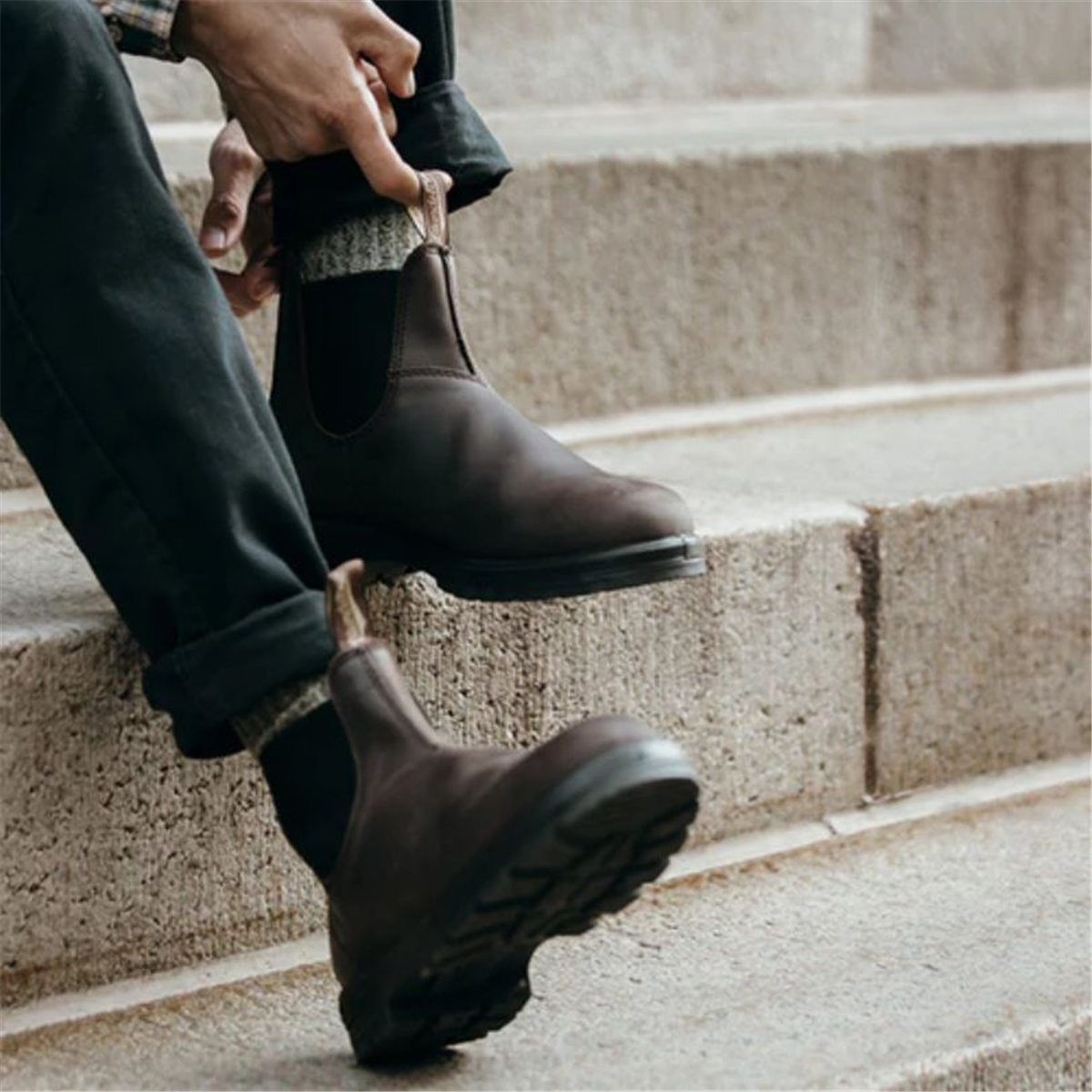 A person sitting on city steps pulling on their brown Blundstone 2340 Classic Series Chelsea boots using the pull tabs.