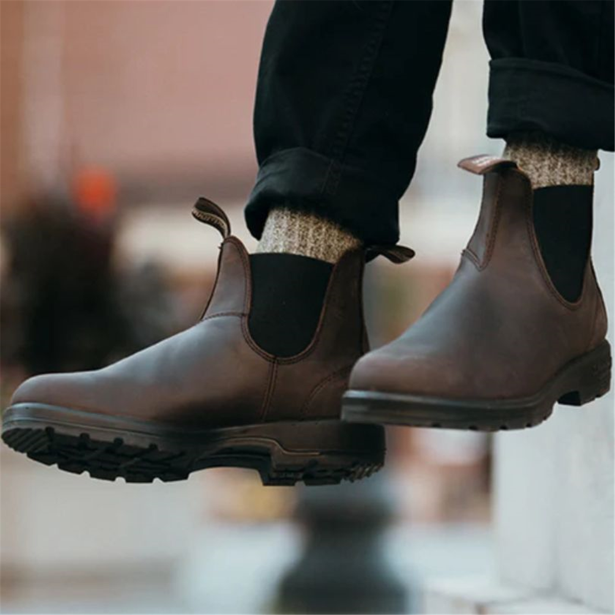 A person sitting with legs dangling, wearing brown Blundstone 2340 Classic Series Chelsea boots against a city backdrop.