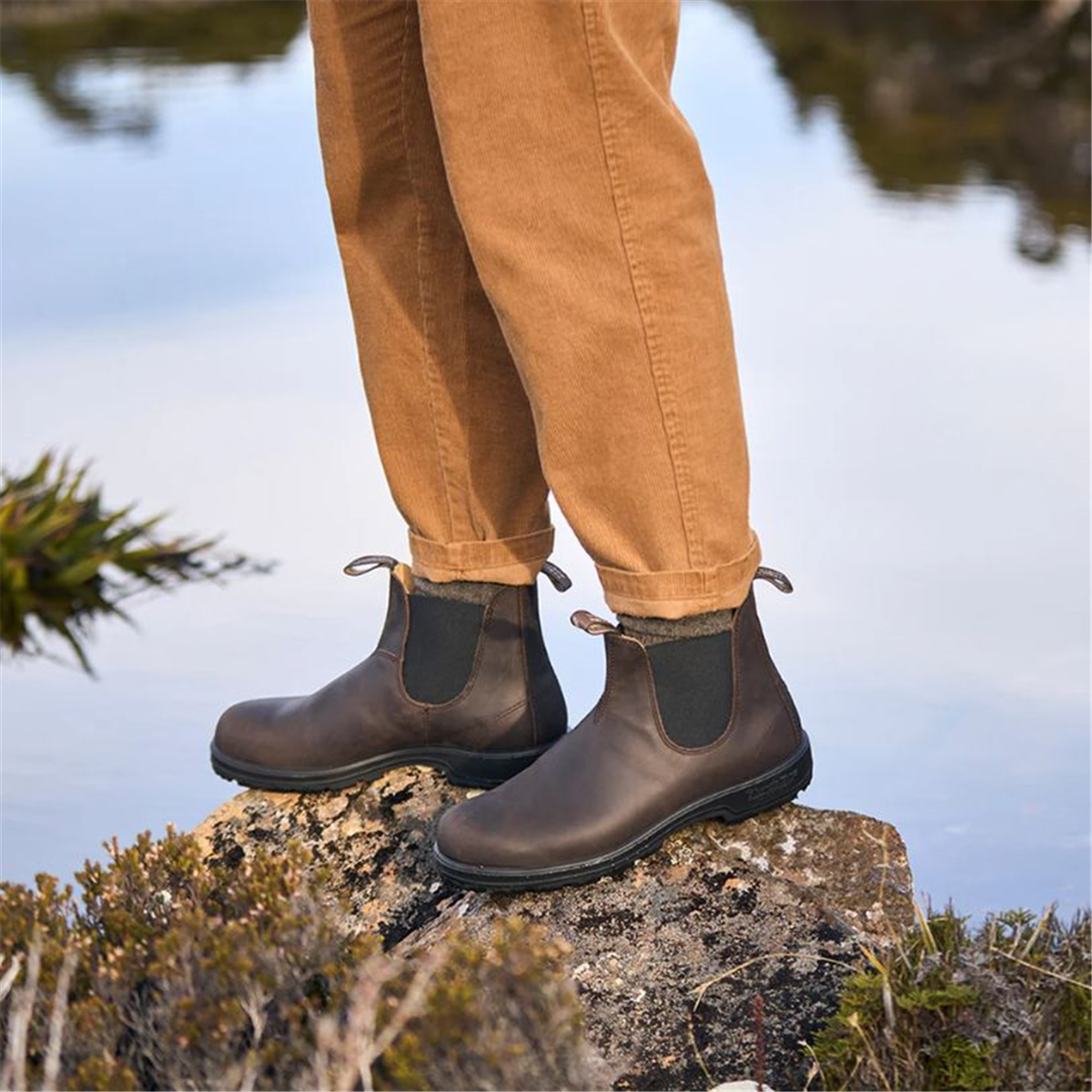 A person standing on a rock next to a serene lake, wearing brown Blundstone 2340 Classic Series Chelsea boots.