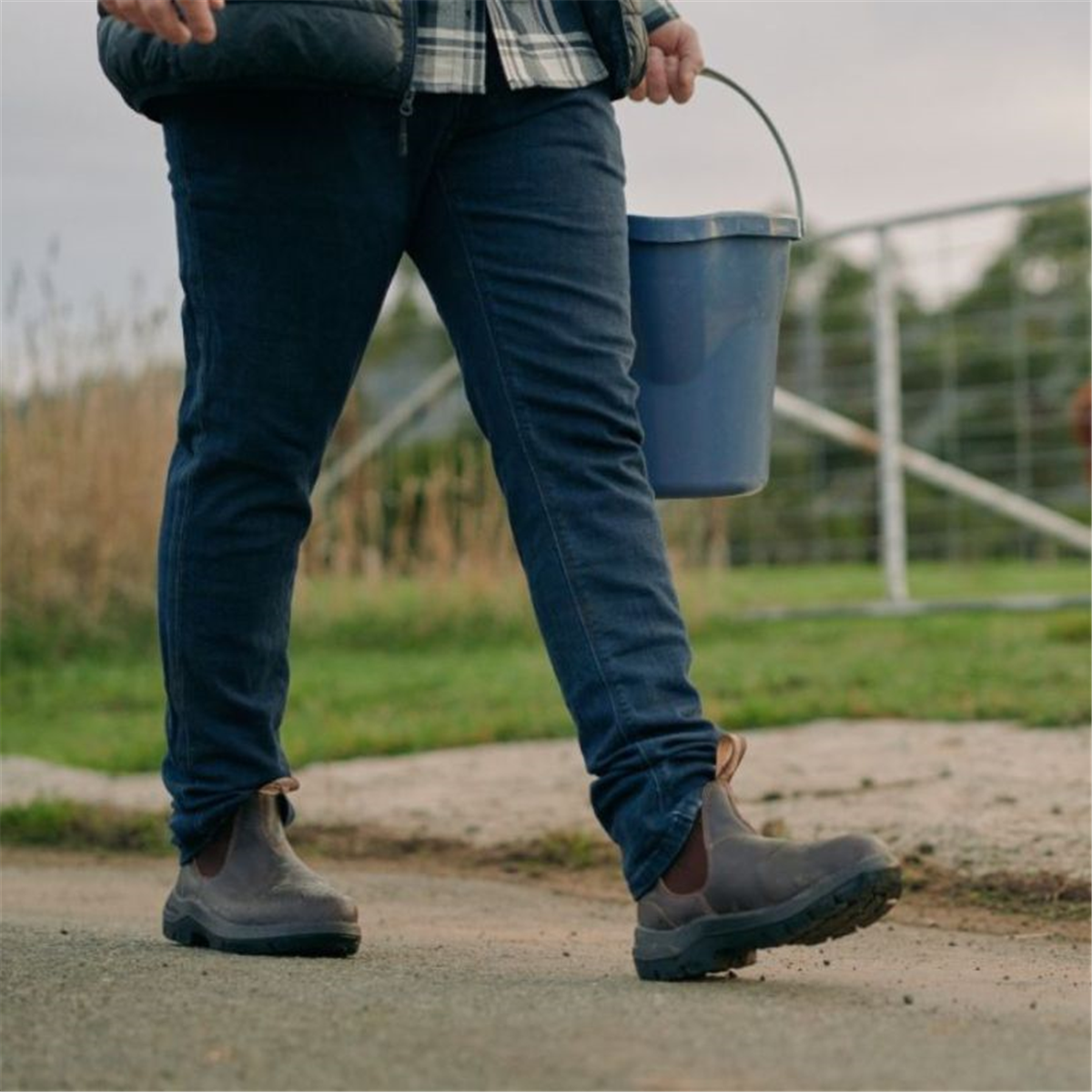Low-angle shot of a person walking outdoors in blue jeans and brown leather Blundstone 710 Chelsea work boots, carrying a blue bucket.
