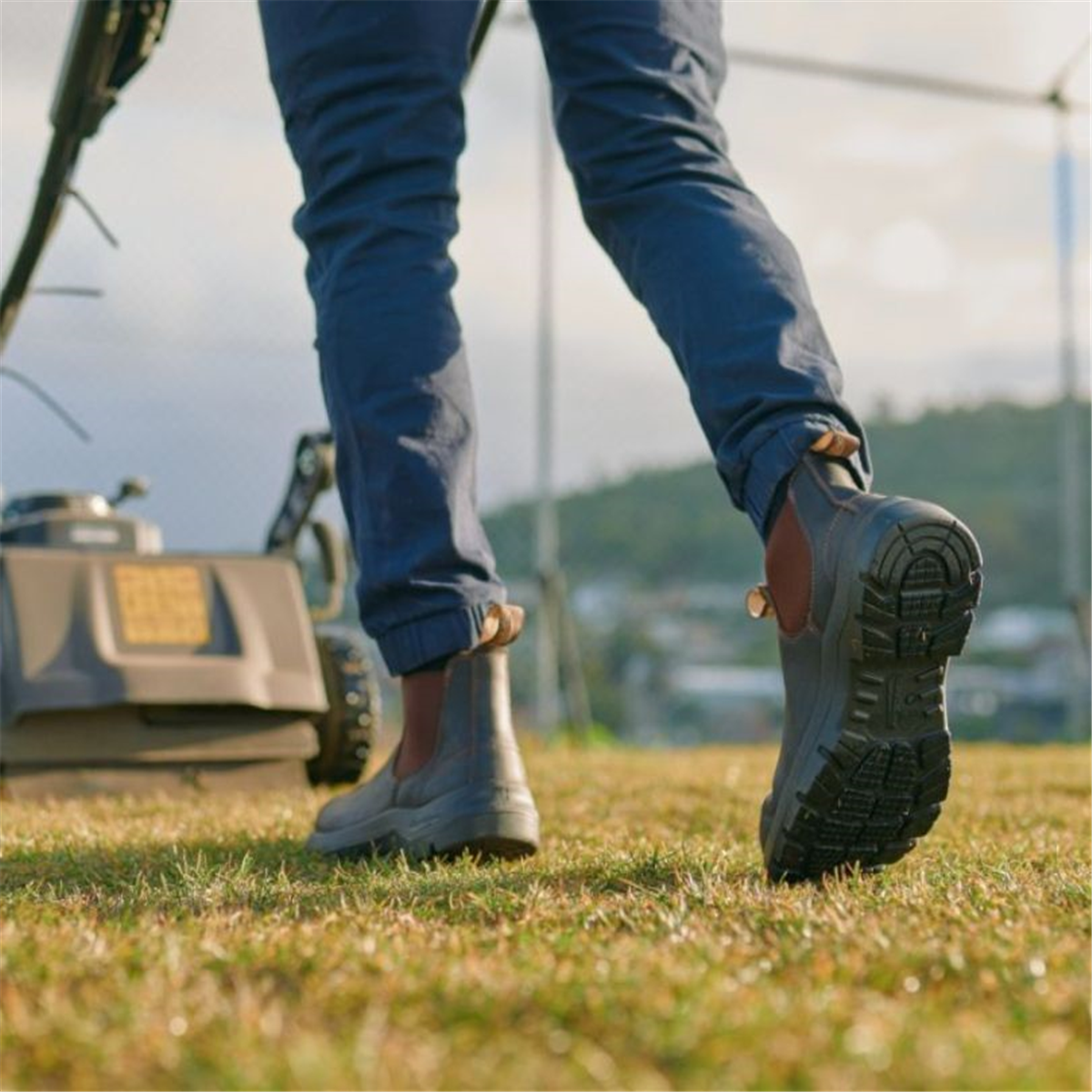 Rear low-angle shot of a person mowing the lawn wearing Blundstone 710 Chelsea work boots, showcasing the thick tread pattern on the raised sole